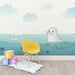 Une chambre d'enfant avec un papier peint illustratif représentant une otarie nageant dans une mer turquoise, entourée de nuages doux et de petits bateaux en papier, créant une atmosphère ludique et apaisante.