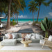 Un salon lumineux avec un papier peint représentant une plage paradisiaque, des palmiers et un chemin en bois menant à une mer turquoise, créant une ambiance relaxante et estivale.